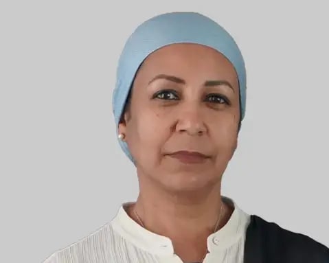 A woman, wearing a light blue head covering and white collared shirt, looks directly at the camera against a plain gray background.