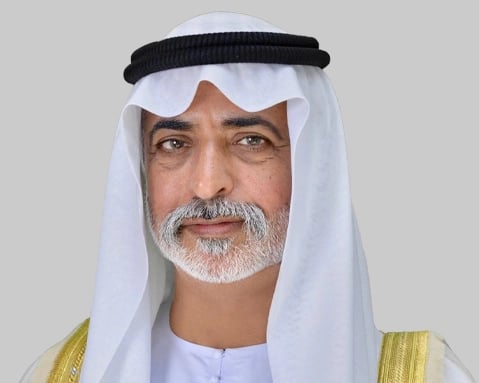 A man, wearing a white head covering and traditional garment, is posing against a light gray background.