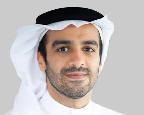 A man, wearing a traditional white thawb and black ghutra, looks directly at the camera against a gray background.
